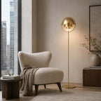Lit beside a cream armchair at a city window, the Clérachic floor lamp in Brass throws warm light down over the seat.