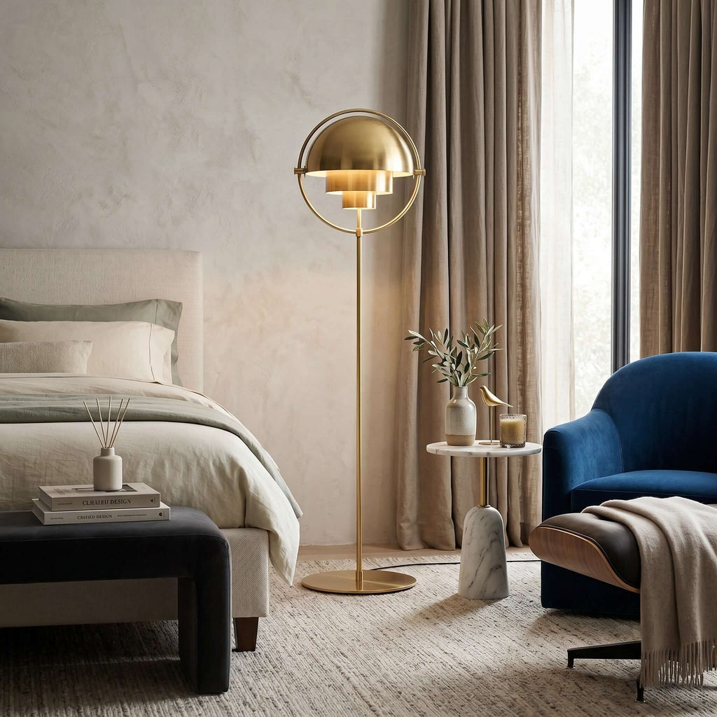 Between a bed and a blue armchair, the lit Clérachic floor lamp in Brass fills the bedroom with a soft, warm glow.