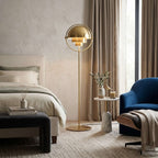 Between a bed and a blue armchair, the lit Clérachic floor lamp in Brass fills the bedroom with a soft, warm glow.