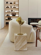A Brass Clérachic table lamp lit on a travertine cube beside a cream bouclé sofa, throwing a warm pool of light across the living room.