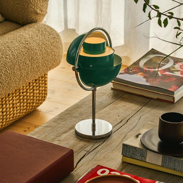 Clérachic lamp in deep green lit on a wooden coffee table beside magazines, shades opened so warm light spills out.