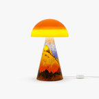 This Clérachic table lamp on a white backdrop, an orange glass dome over a cone base painted with golden hills, with a cord switch on the cable.