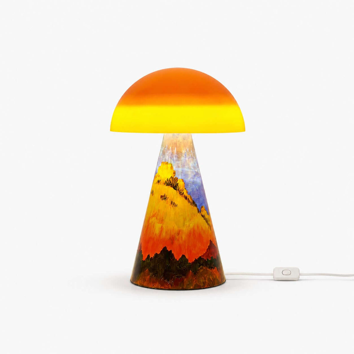 This Clérachic table lamp on a white backdrop, an orange glass dome over a cone base painted with golden hills, with a cord switch on the cable.