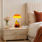 Lit Clérachic table lamp on a cream bedside chest with two drawers, beside pink flowers, a tray and a bed with rust cushions.