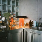 Orange-based Clérachic table lamp with a dark shade, on a steel kitchen counter in front of a glass-block wall.