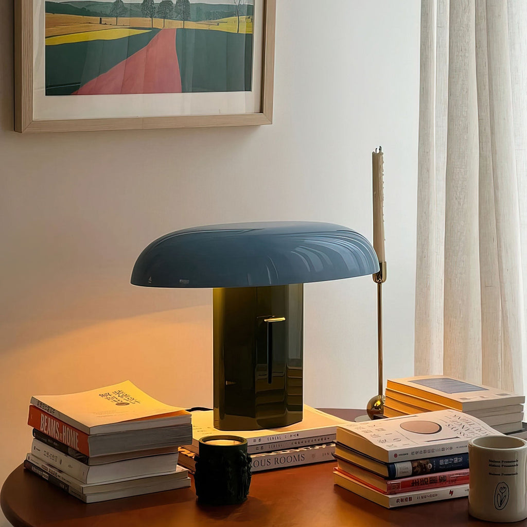 Lit Clérachic table lamp with a blue dome shade and olive-green base, glowing warmly on a wooden table beside stacked books.