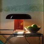 Clérachic table lamp with a dark shade and red-orange column, on a glass side table beside a bowl of green apples.