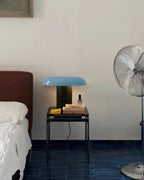 This Clérachic table lamp with a blue shade stands on a small black side table beside a bed, with a metal fan nearby.