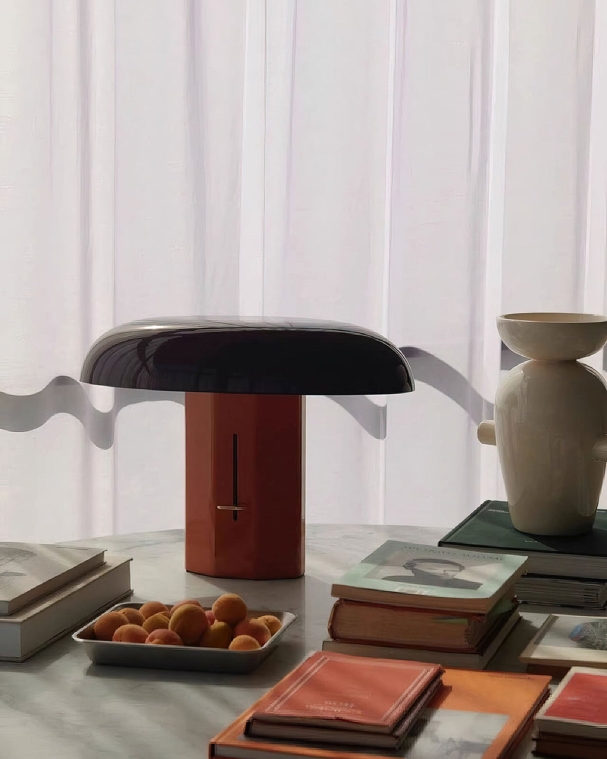 Red-orange base and dark domed shade on a Clérachic table lamp, among books and a bowl of apricots on a marble surface.