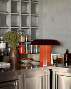 Clérachic table lamp in orange and dark burgundy on a steel counter, with a dried-flower vase and glass jars behind.