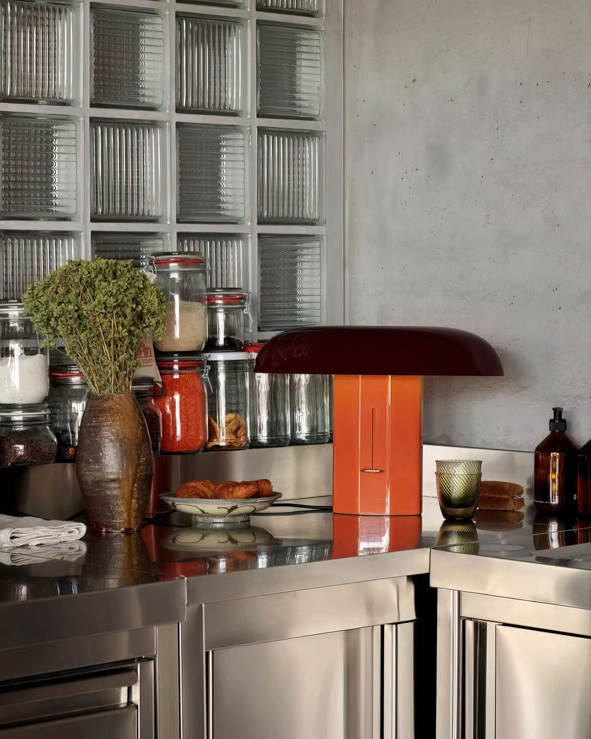 Clérachic table lamp in orange and dark burgundy on a steel counter, with a dried-flower vase and glass jars behind.
