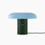 Front view of a Clérachic table lamp with a pale blue shade and green base, its black cord trailing to the right.