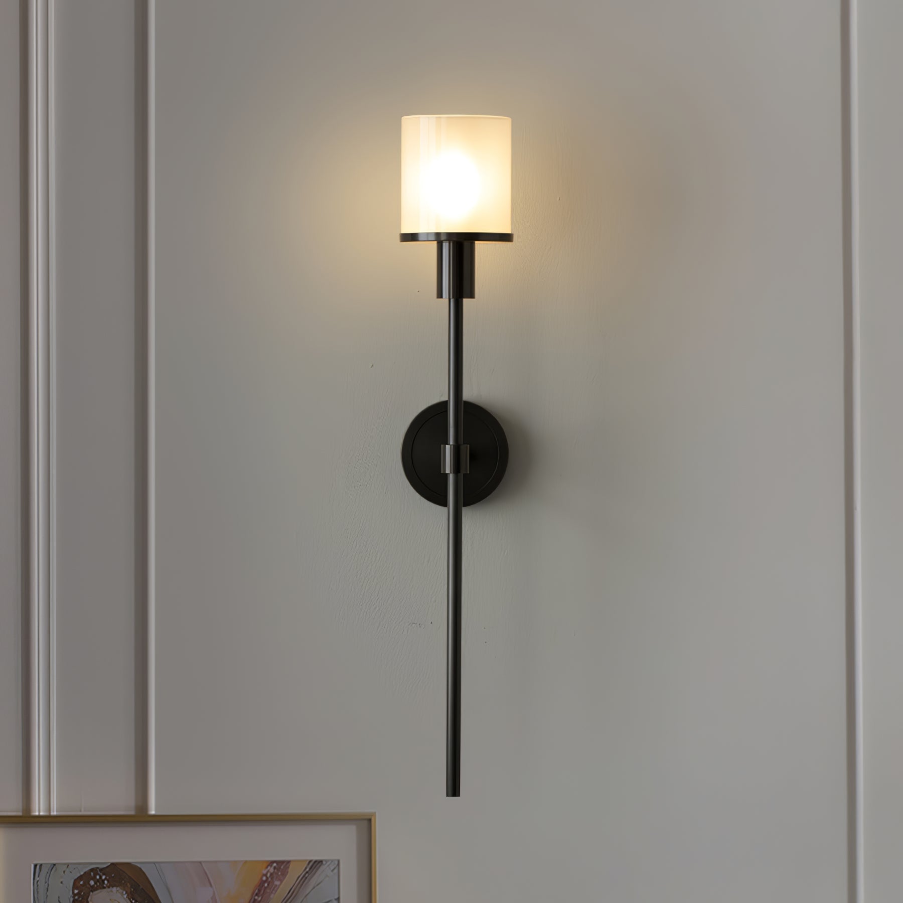 The Clérachic Mycara in black, lit on a pale panelled wall above a framed print resting at the skirting.