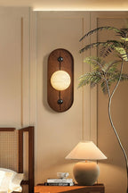 The Clérachic sconce glows beside a bed, next to a slim indoor palm and a ceramic table lamp on the side table.