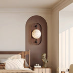 Clérachic sets the sconce on a deep brown arch, lighting a bedroom alcove beside a cane headboard and linen bedding.