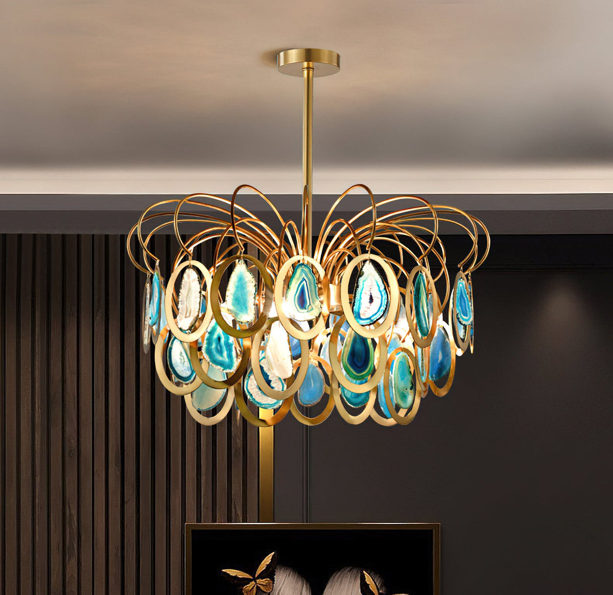 A Clérachic chandelier in blue agate and gold hung before a dark slatted wall, with a framed picture just visible below.