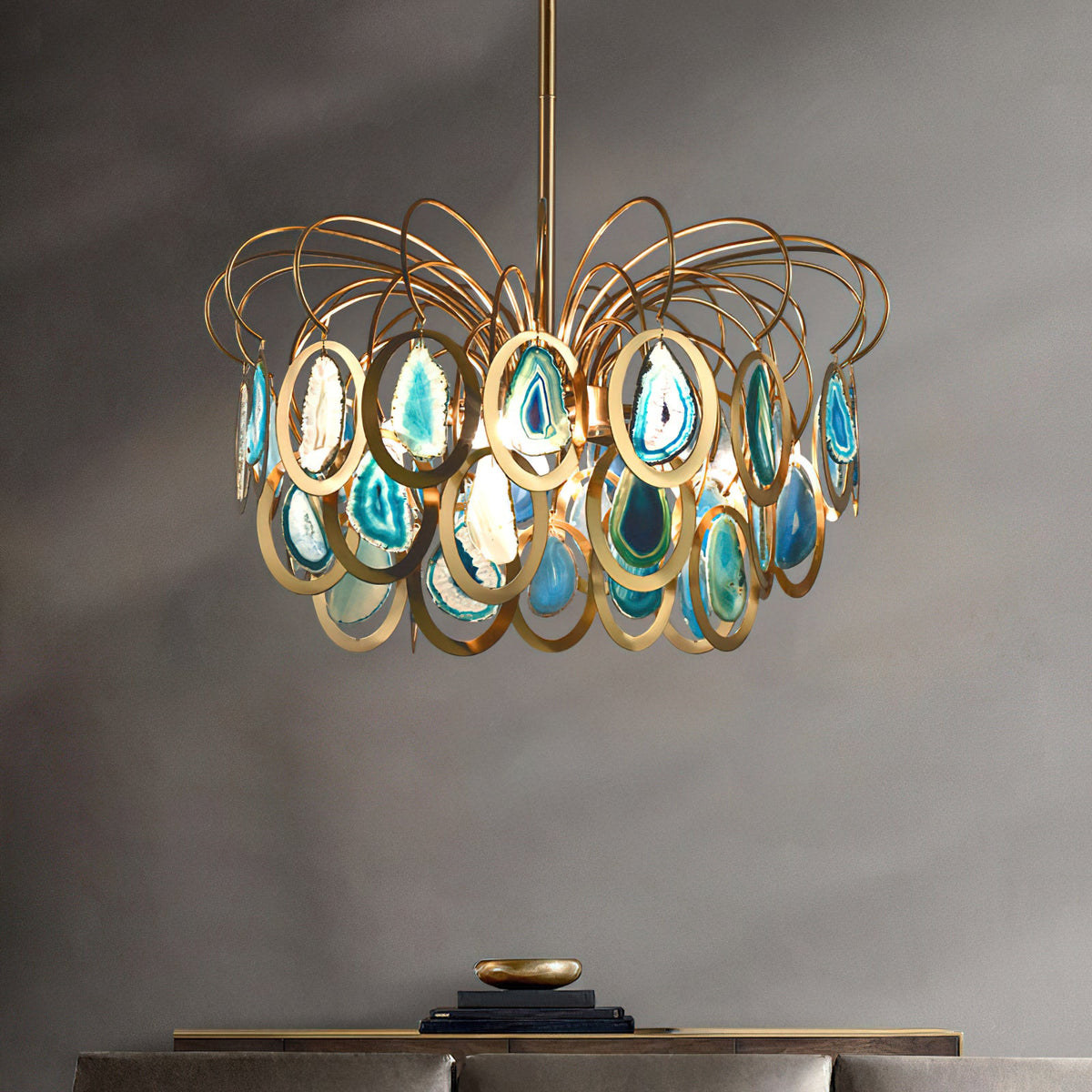 Blue and gold Clérachic chandelier hanging in front of a grey wall, a console with a brass bowl and books below it.