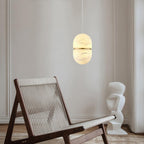 This Clérachic alabaster pendant glowing warm in a pale room, above a rope-seat lounge chair and a white sculptural object.