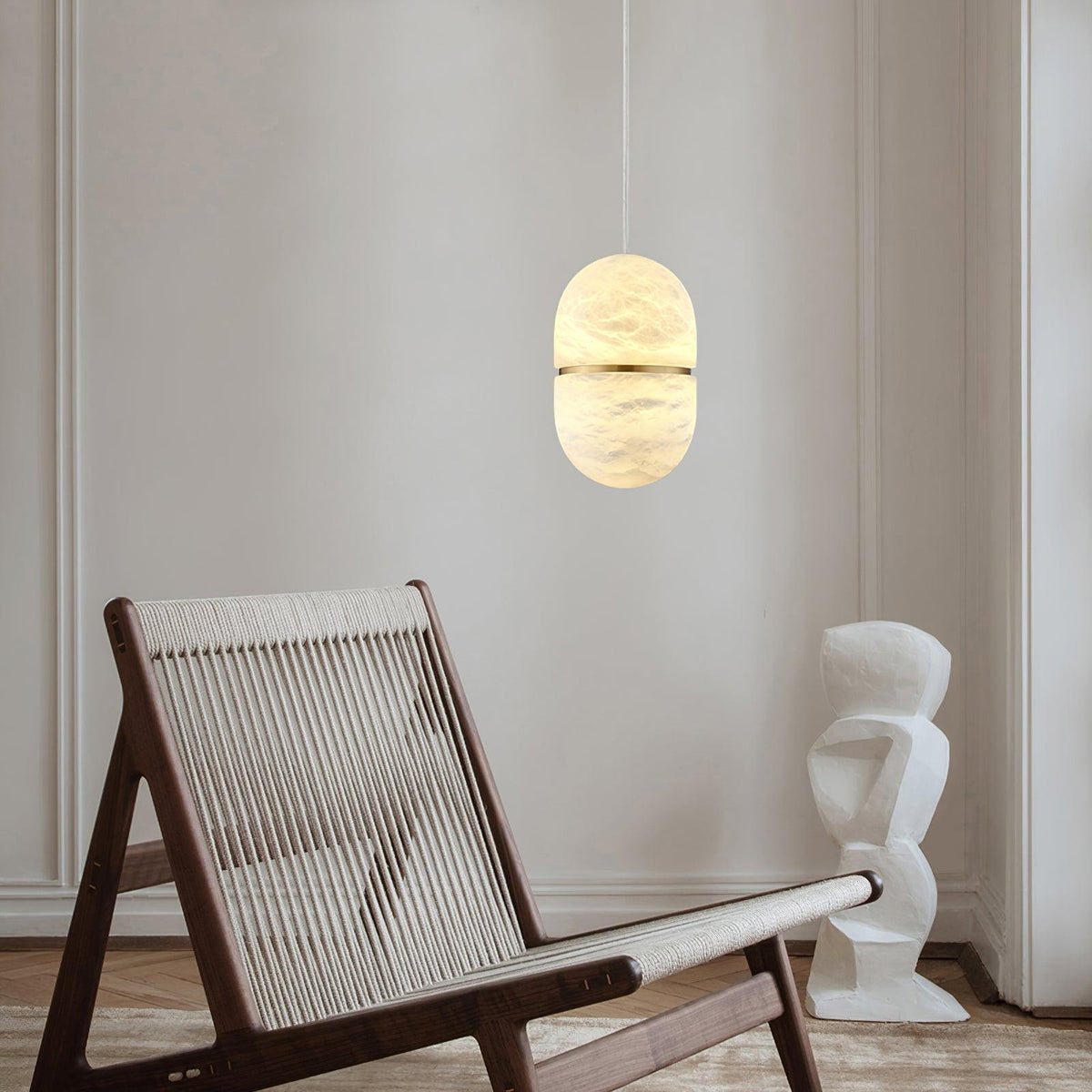This Clérachic alabaster pendant glowing warm in a pale room, above a rope-seat lounge chair and a white sculptural object.