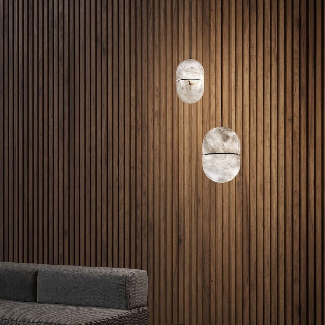Two Clérachic alabaster pendants against dark vertical timber slats, with the edge of a grey sofa at the bottom of the frame.