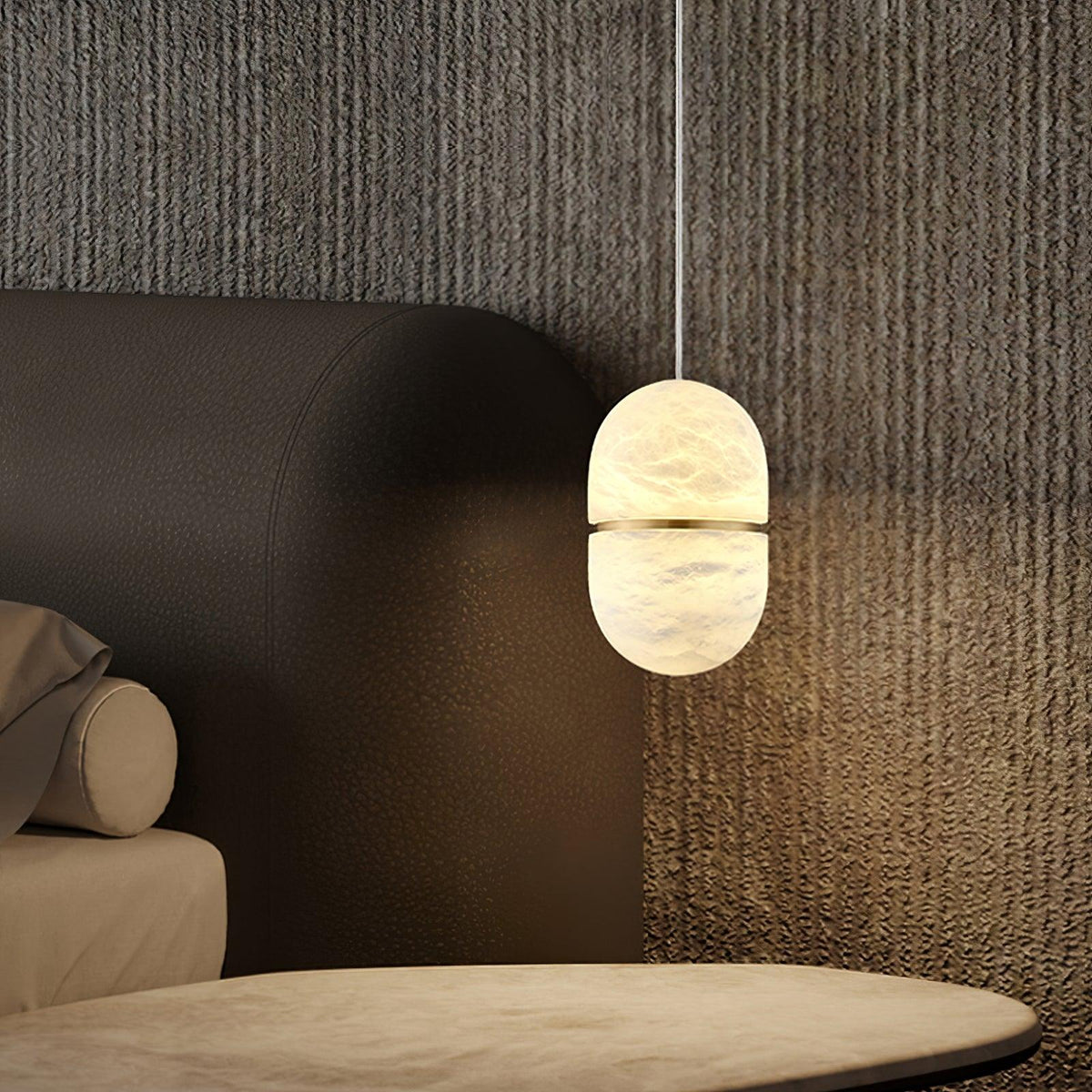 A lit Clérachic alabaster pendant beside a dark leather headboard and a stone-topped table, against a textured grey wall.