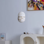 This Clérachic alabaster pendant, unlit, against a pale lilac wall with a colourful abstract painting and a white arched console below.