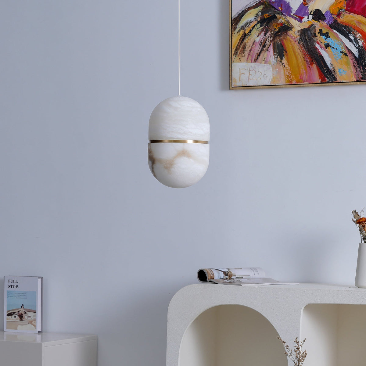 This Clérachic alabaster pendant, unlit, against a pale lilac wall with a colourful abstract painting and a white arched console below.