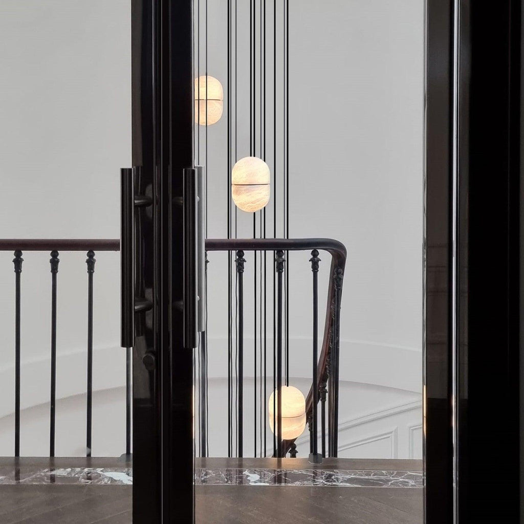 Clérachic alabaster pendants hung at different heights beside a black-framed doorway and a stair railing in a pale hall.
