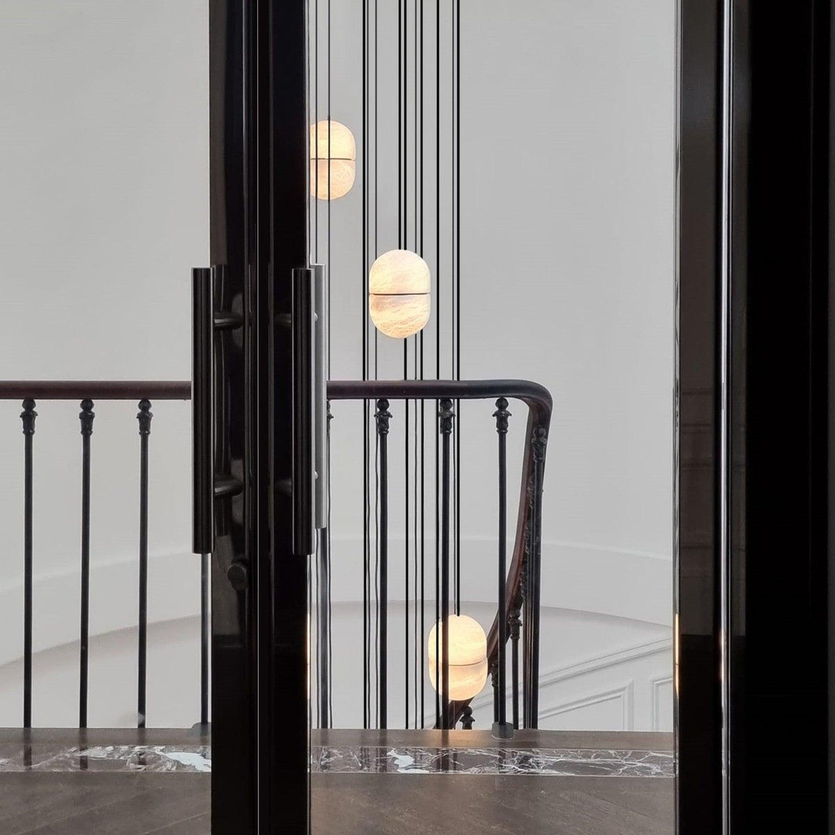 Clérachic alabaster pendants hung at different heights beside a black-framed doorway and a stair railing in a pale hall.