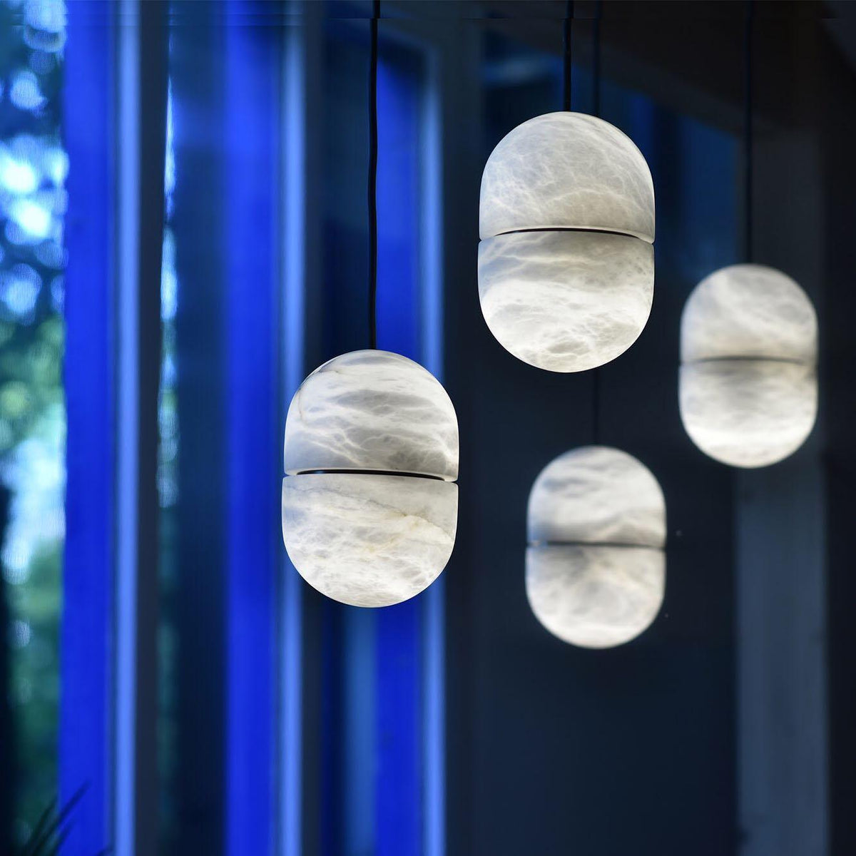 Four Clérachic alabaster pendants glowing at different heights against a dark window with blue light behind.
