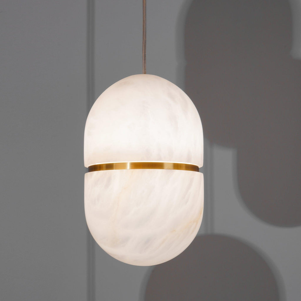 A Clérachic alabaster pendant with a gold band, glowing against a grey wall with soft shadows of other shades behind it.
