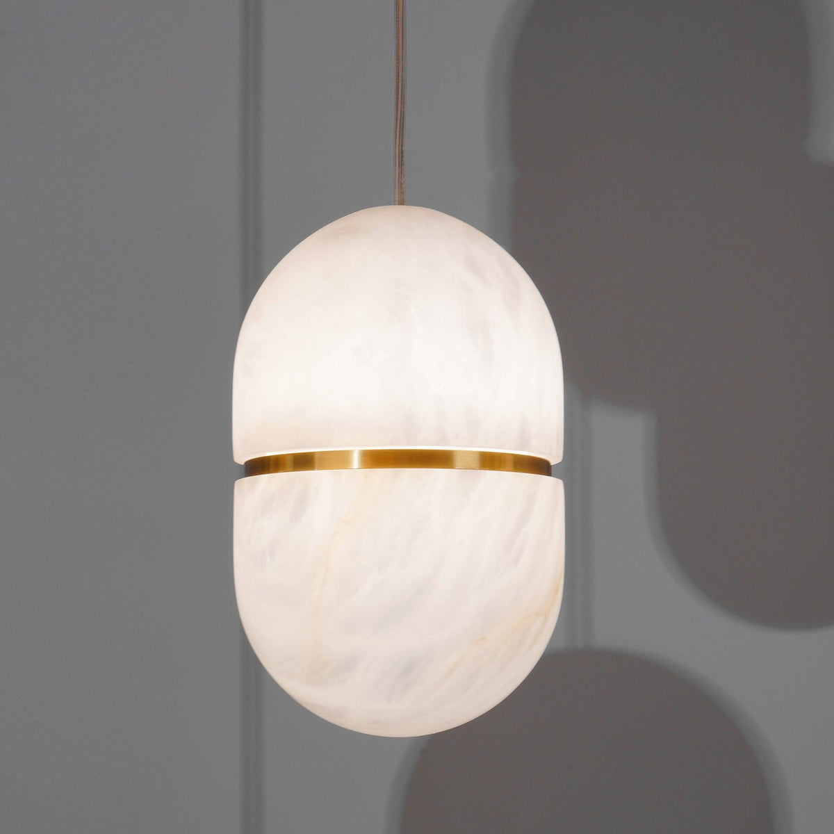 A Clérachic alabaster pendant with a gold band, glowing against a grey wall with soft shadows of other shades behind it.
