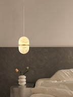 A small Clérachic alabaster pendant on a cord above a bedside table with a stacked white vase, beside a rumpled bed.