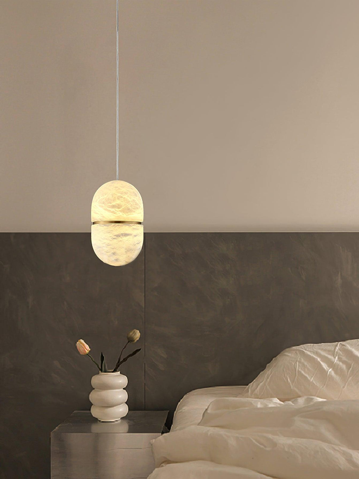 A small Clérachic alabaster pendant on a cord above a bedside table with a stacked white vase, beside a rumpled bed.