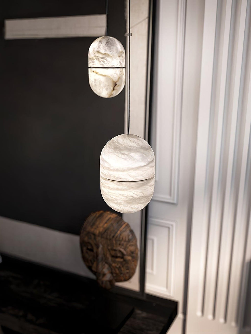Two Clérachic alabaster pendants hung one above the other in a dark corner, above a carved wooden mask and a white fluted wall.