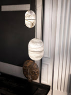 Two Clérachic alabaster pendants hung one above the other in a dark corner, above a carved wooden mask and a white fluted wall.