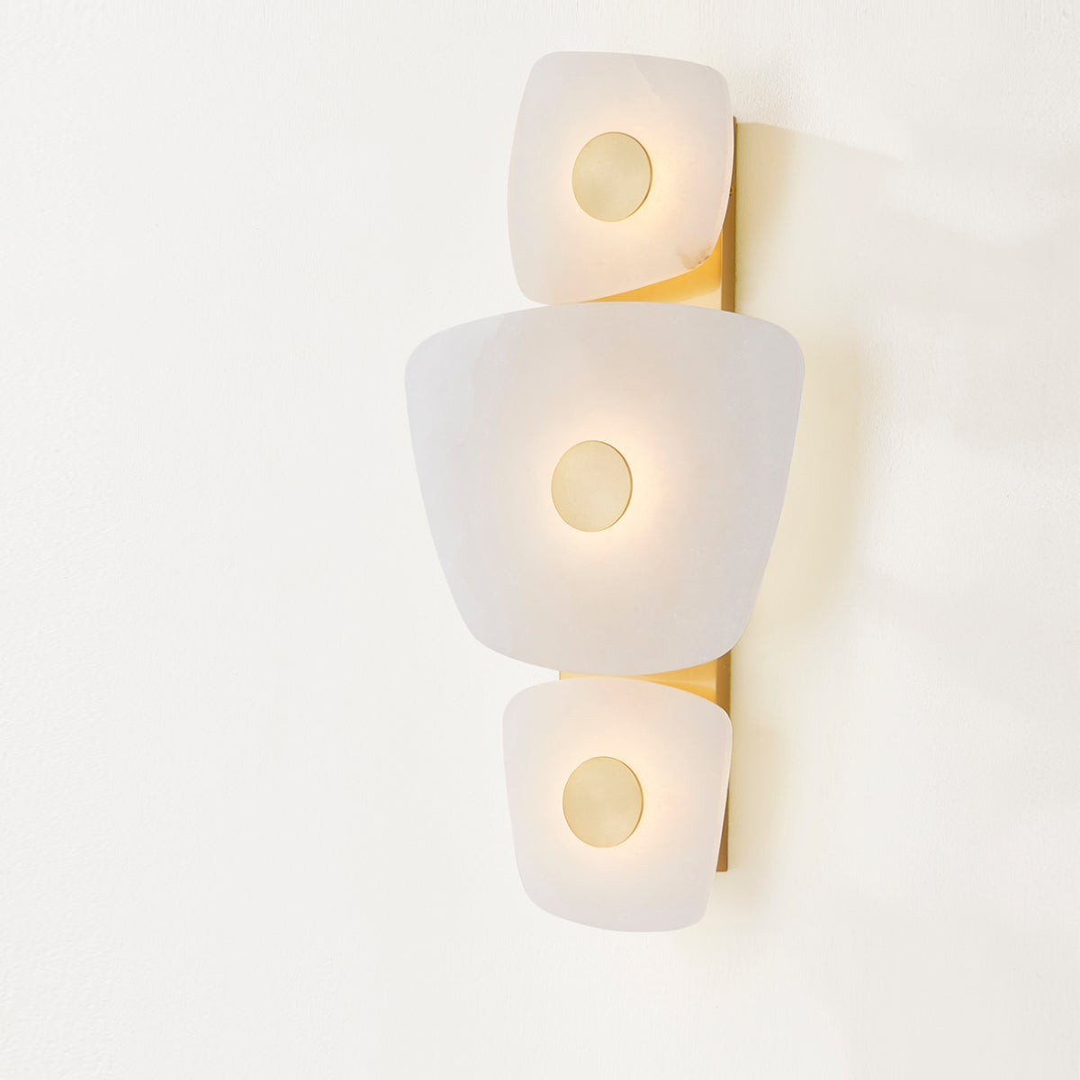 Clérachic alabaster wall sconce lit on a plain wall, the slim brass backplate visible behind the three glowing plates.