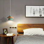 A green-shaded Clérachic pendant with a wood top hangs beside a wooden headboard, below a framed feather print on a grey wall.