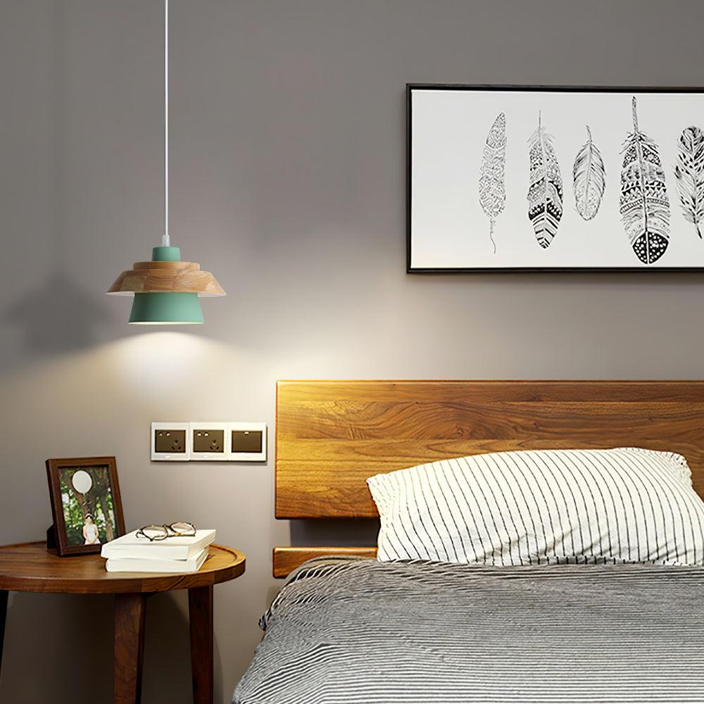 A green-shaded Clérachic pendant with a wood top hangs beside a wooden headboard, below a framed feather print on a grey wall.