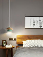 A green-shaded Clérachic pendant with a wood top hangs near a bed, with a feather print and a round wooden side table.
