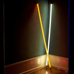 Two Clérachic neon-style LED floor tubes, one yellow and one white, crossed in an X against a dark green corner wall with their caps resting on a wood floor.