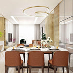 This Clérachic gold ring chandelier curls above a long dining table with rust-brown chairs, in a bright room with timber slats.