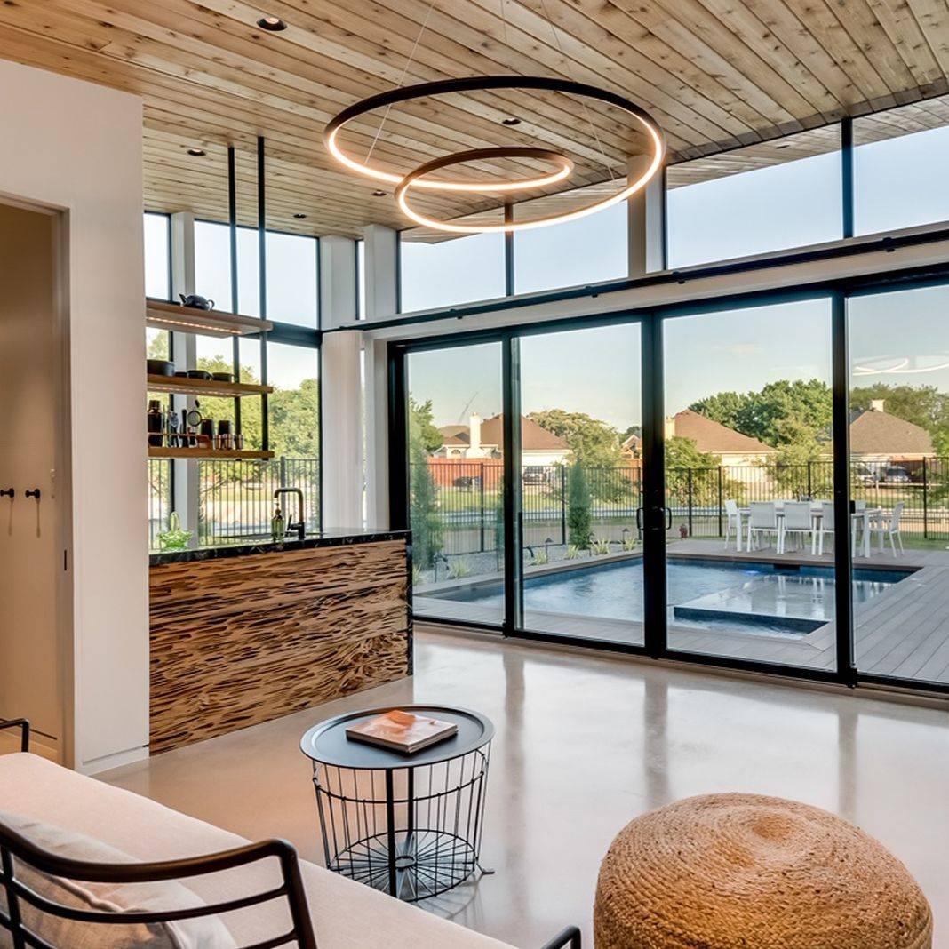 Gold double-ring Clérachic chandelier hung from a timber ceiling above an open living space with glass doors and a pool beyond.