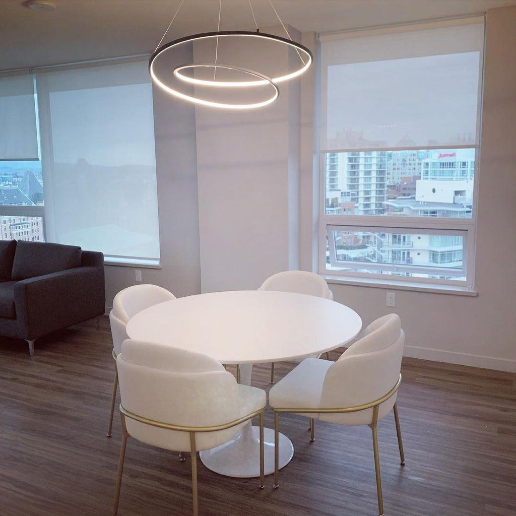 Black nested-ring Clérachic chandelier lit above a round white table and cream chairs, by tall windows with roller blinds.