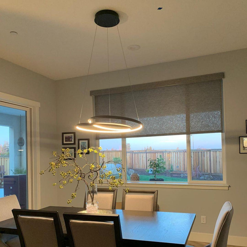 A Clérachic black ring chandelier lit above a dark dining table, in a room with a window blind and a vase of yellow blossom.