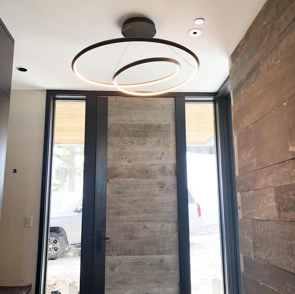 Black loop chandelier from Clérachic hung in a tall entrance hall, with a weathered wood door and a dark plank wall.