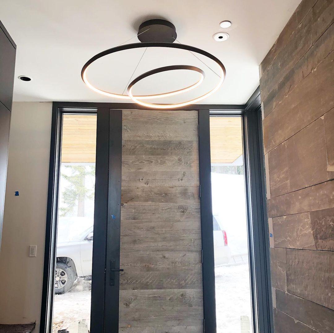 Black loop chandelier from Clérachic hung in a tall entrance hall, with a weathered wood door and a dark plank wall.