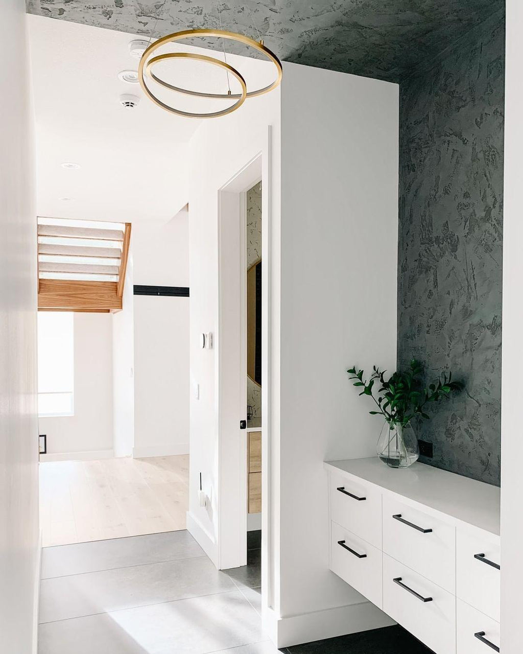 Small gold ring chandelier from Clérachic in a white hallway beside a dark textured wall and a white sideboard.