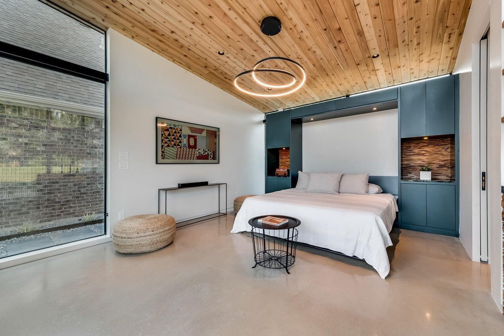Black ceiling-mounted Clérachic ring chandelier lit above a bed with a pale cedar ceiling, between a teal cabinet and a rattan pouffe.