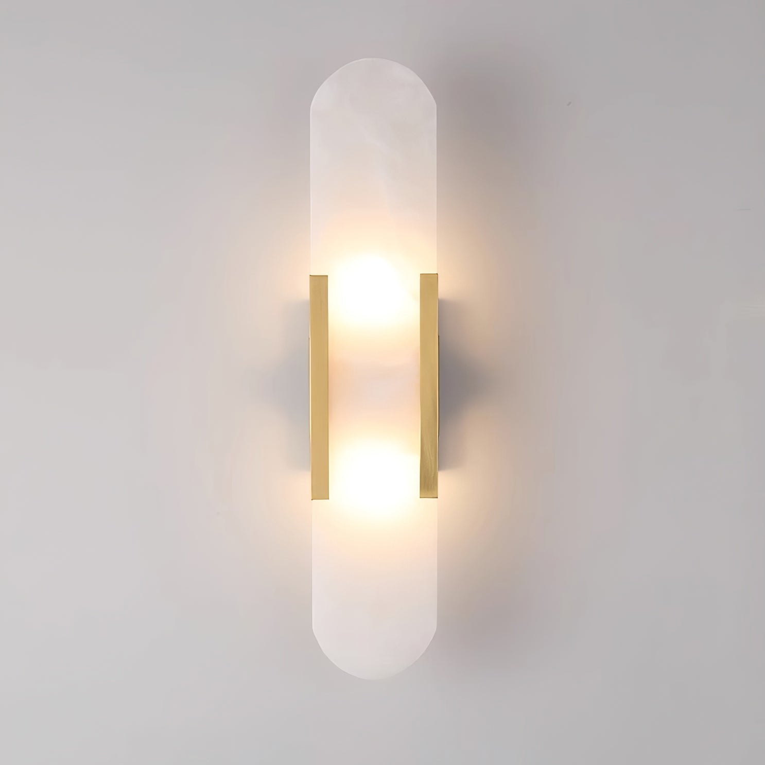 Clérachic Olraeia wall sconce in brass, both bulbs glowing behind the capsule alabaster panel on a pale grey wall